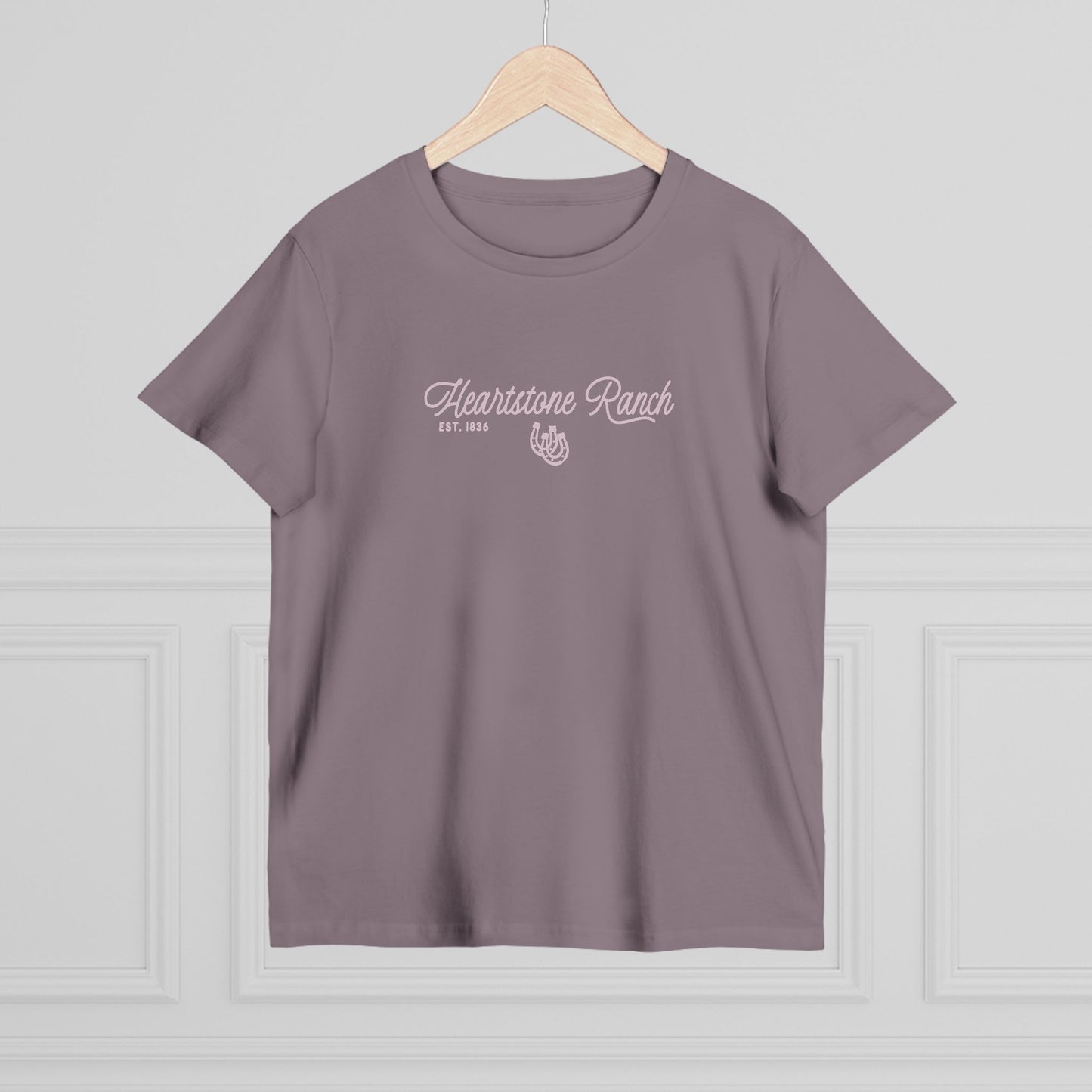 A mauve-purple short-sleeve t-shirt hanging on a light wood hanger against a white paneled wall. The shirt features a white screen-printed graphic on the chest reading 'Heartstone Ranch EST.