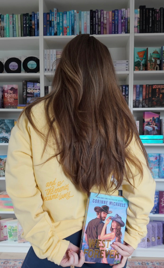 A woman with long brown hair, seen from behind, wearing a pale yellow hoodie and holding a book titled 'West Forever' by Corinne Michaels in front of a bookshelf filled with books.