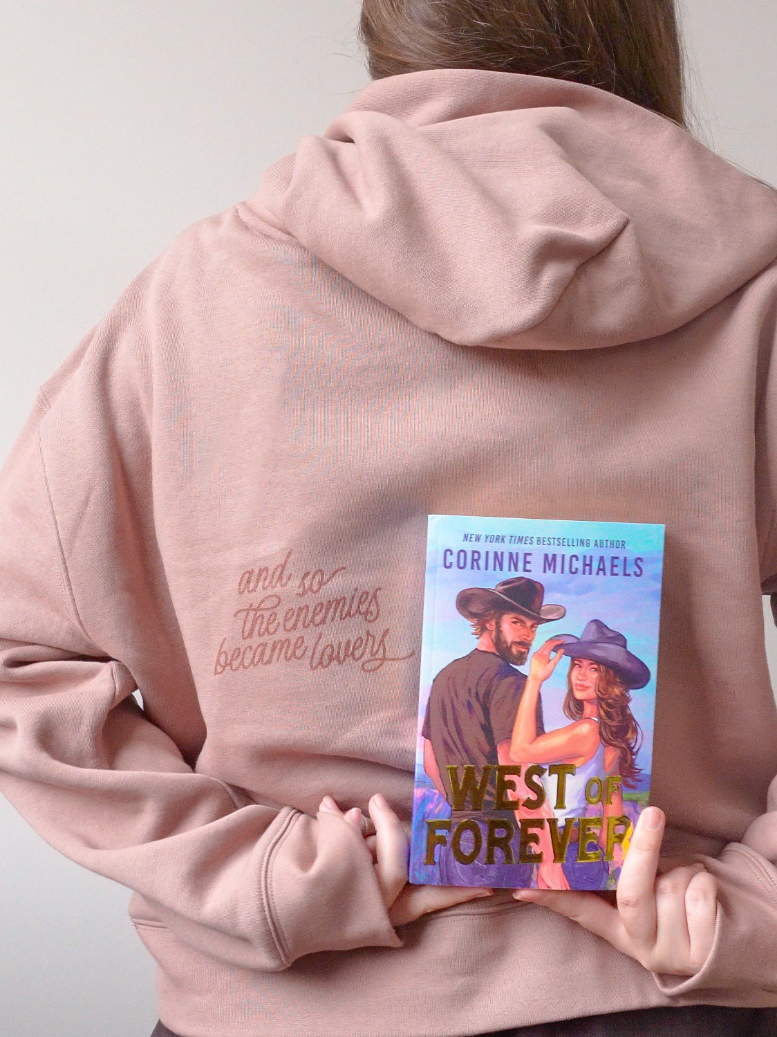 A person wearing a dusty pink hoodie with gold cursive embroidery on the back, holding a book titled 'West of Forever' by Corinne Michaels in front of their back.