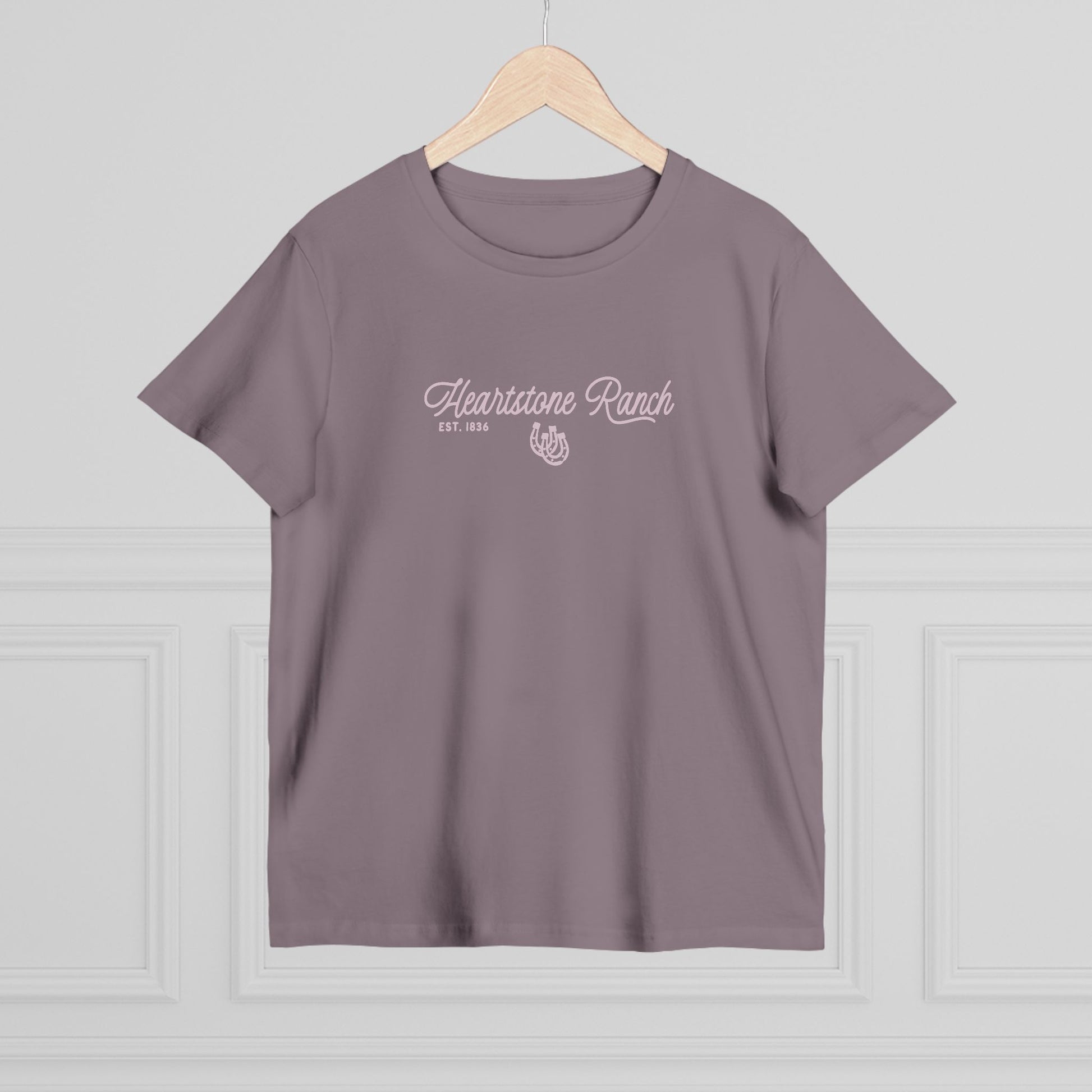 A mauve-purple short-sleeve t-shirt hanging on a light wood hanger against a white paneled wall. The shirt features a white screen-printed graphic on the chest reading 'Heartstone Ranch EST.