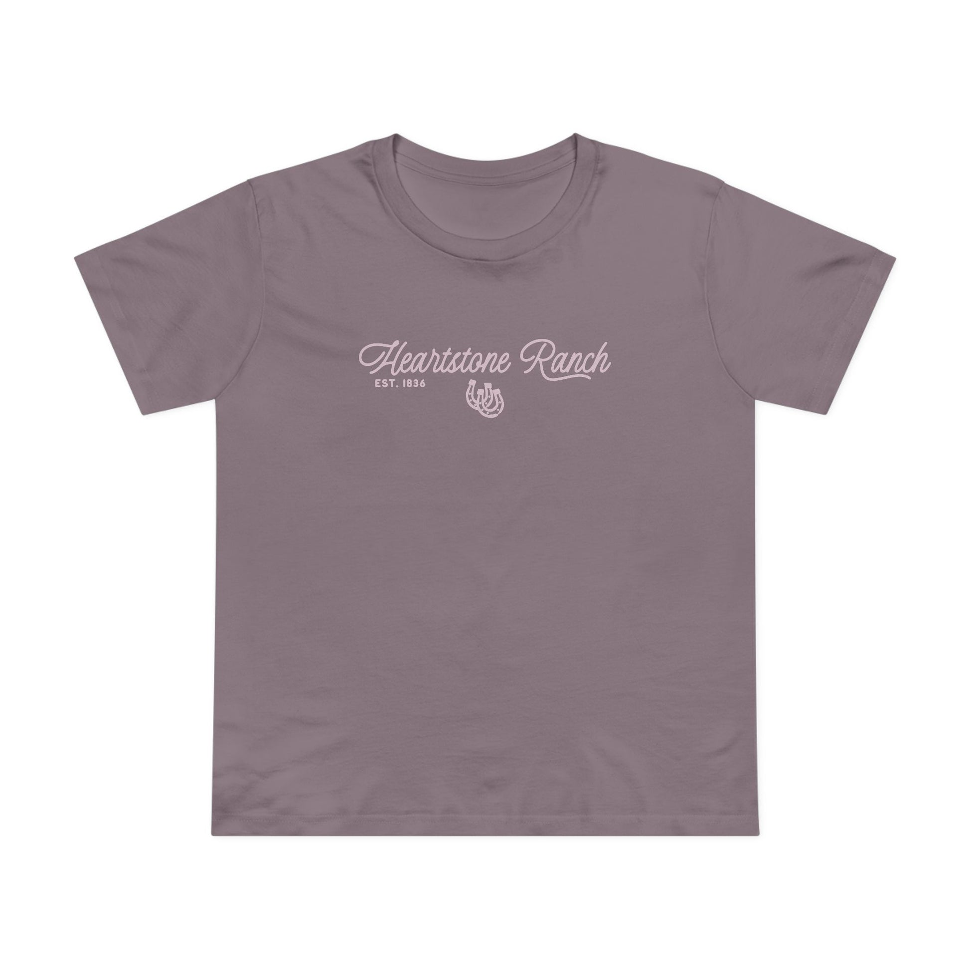A mauve-purple short-sleeve t-shirt hanging on a light wood hanger against a white paneled wall. The shirt features a white screen-printed graphic on the chest reading 'Heartstone Ranch EST.