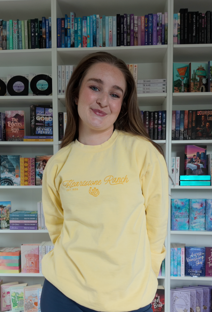 A young woman with long brown hair stands in front of white bookshelves filled with books, wearing a pale yellow long-sleeve crewneck sweatshirt with 'Heartstone Ranch' printed in gold script across the chest.