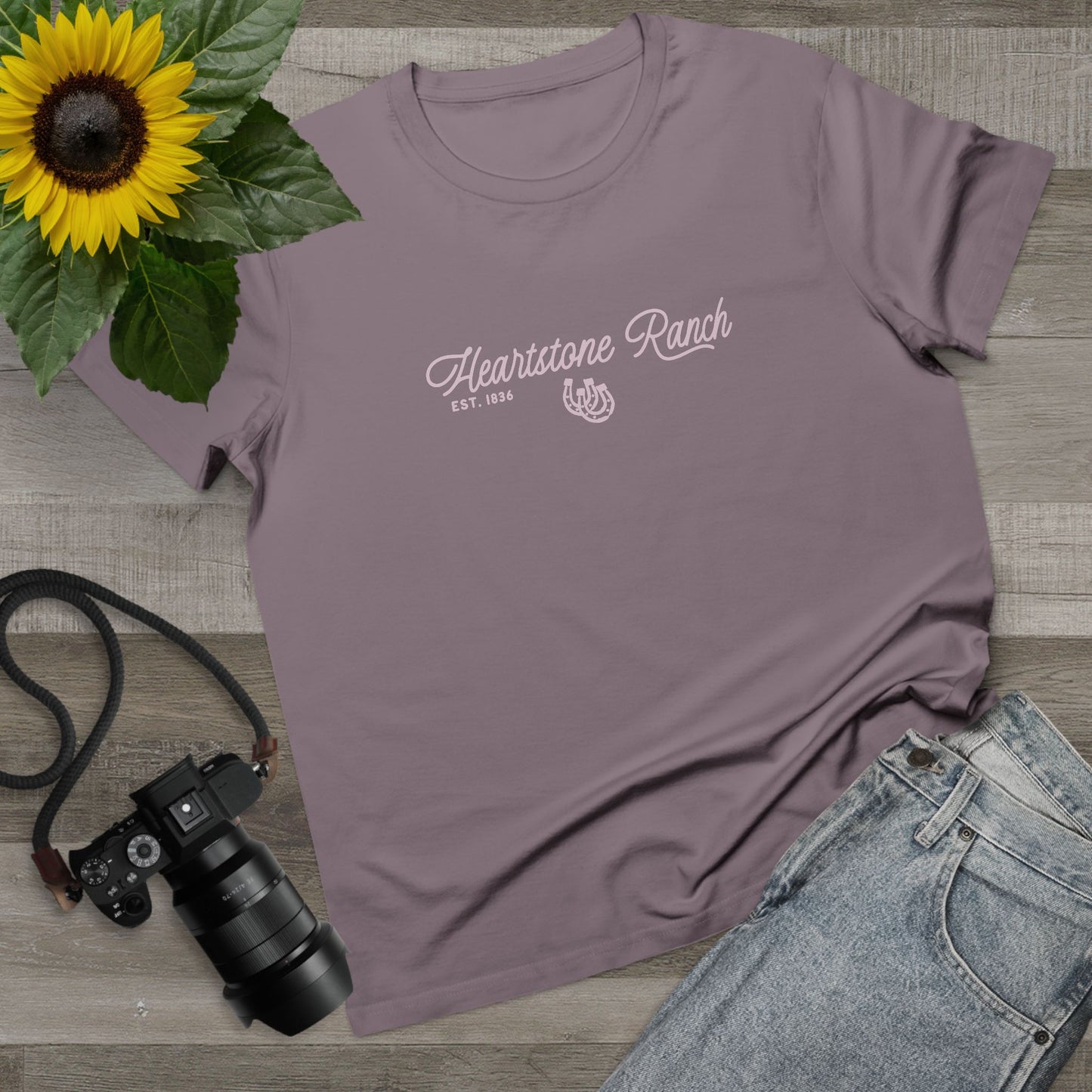 A mauve-purple short-sleeve t-shirt with 'Heartstone Ranch EST. 1865' printed in white script on the chest, styled in a flat lay with a sunflower, camera, and denim jeans on a wood background.
