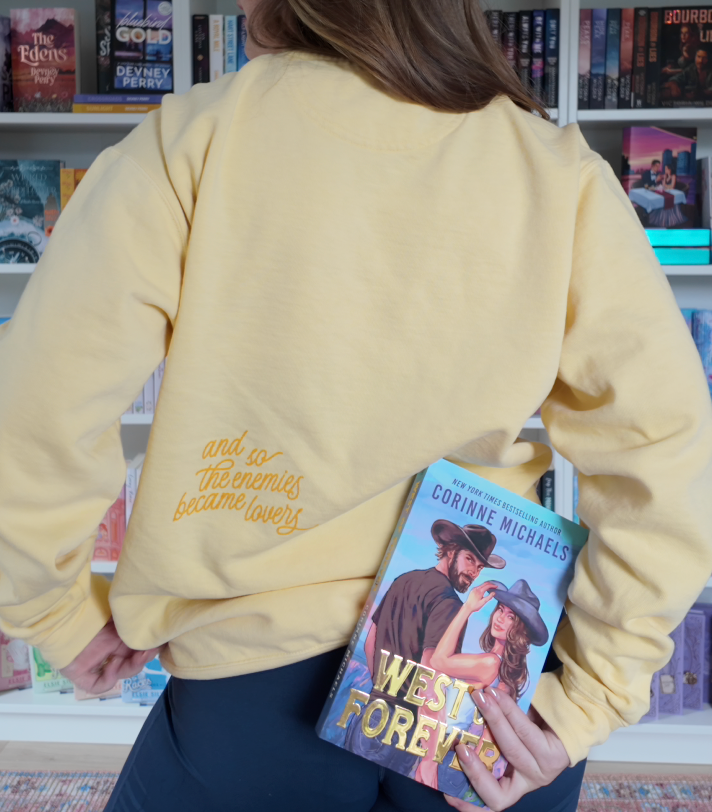 A person wearing a pale yellow sweatshirt stands in front of bookshelves, holding a book under their arm. The sweatshirt has gold cursive text on the back that reads 'and so the enemies become lovers.