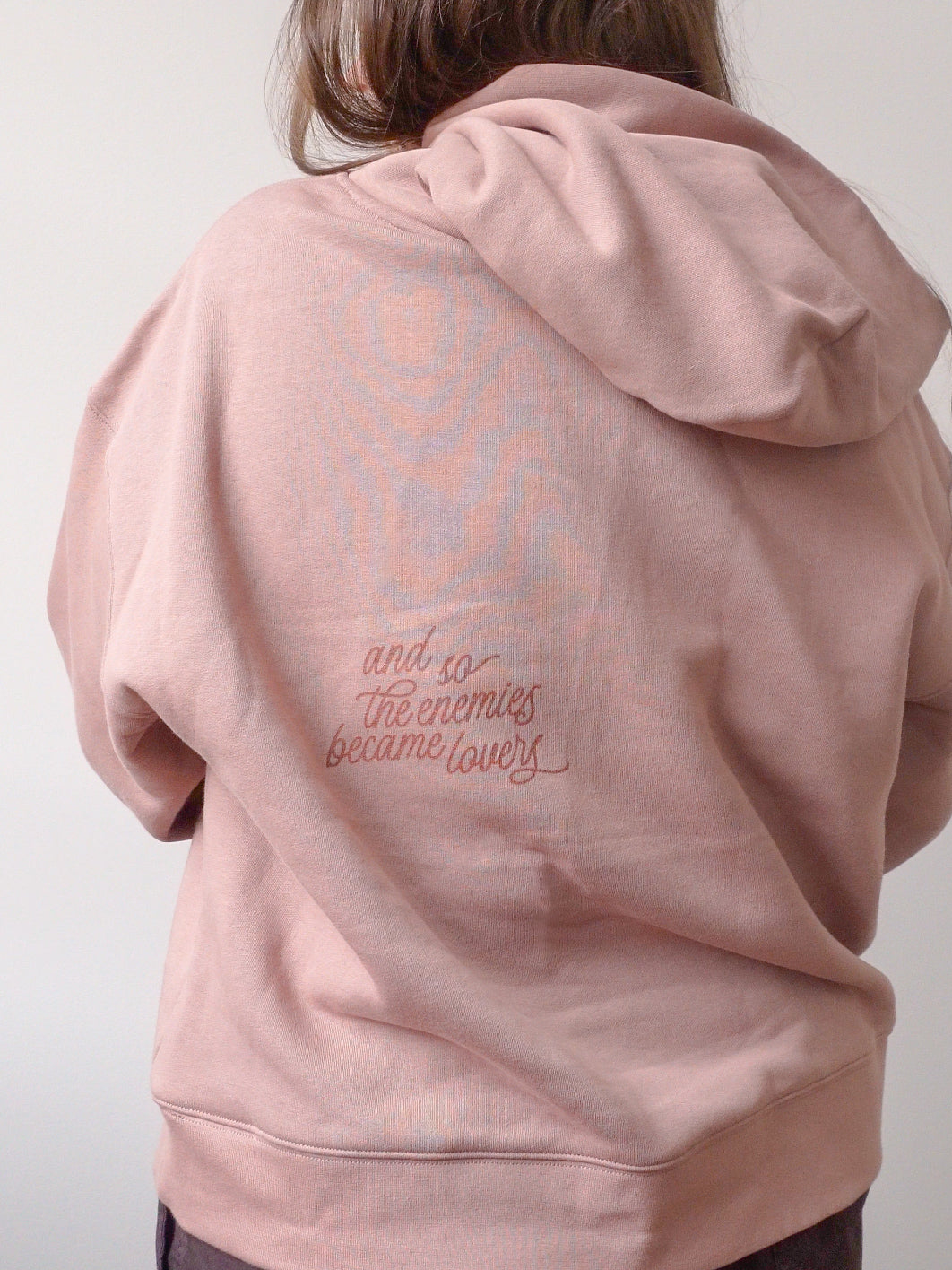 A person viewed from behind wearing a dusty pink hoodie with a faint rose graphic and the embroidered phrase 'and so the enemies became lovers' on the back.