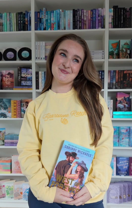 A young woman with long light brown hair stands in front of white bookshelves filled with books, wearing a pale yellow sweatshirt with gold script text and holding a book titled 'West of Forever' by Corinne Michaels.