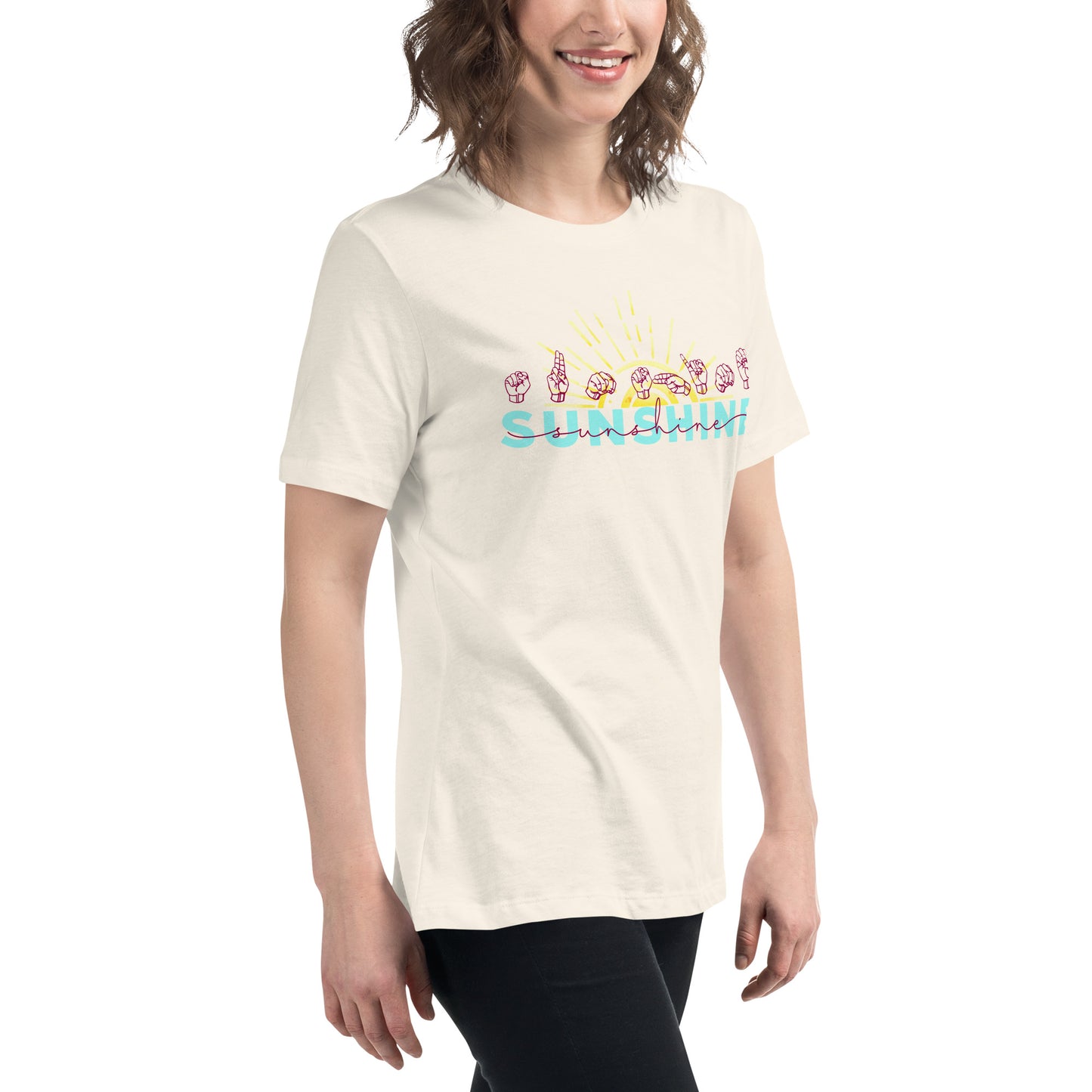 A woman wearing a cream-colored t-shirt with a colorful graphic that reads 'SUNSHINE' with hand signs and a sun, paired with black pants, against a plain white background.